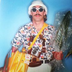 a man with sunglasses and a hat holding a yellow bag in his right hand while standing next to a blue wall