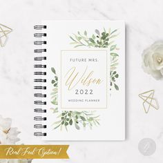 a white wedding planner with greenery and gold foil lettering on it, next to flowers