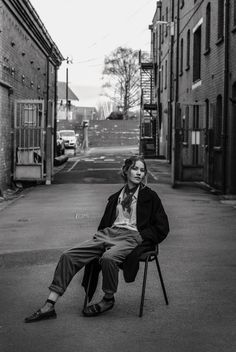 black and white photograph of woman sitting on chair in alleyway with buildings behind her