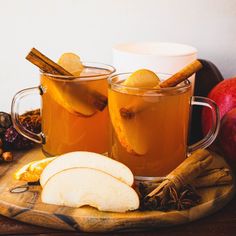 two mugs filled with apple cider on top of a wooden tray next to apples and cinnamon sticks