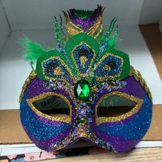 a purple mask with green feathers and a jewel in the center, sitting on top of a cardboard box