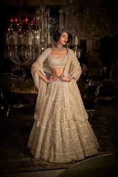 a woman in a white and gold lehenga standing next to a chandelier