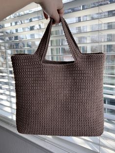 a hand holding a brown crocheted bag in front of a window with blinds