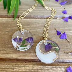 two necklaces with flowers in them sitting on a wooden table next to purple flowers