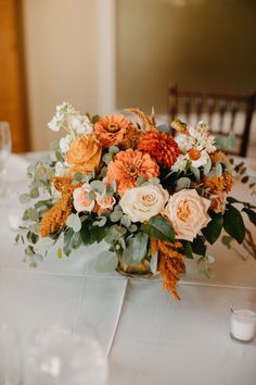 an arrangement of flowers in a vase on a white table cloth with candles and glasses