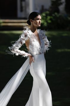 a woman wearing a white dress and jacket with feathers on her arms, standing in the grass