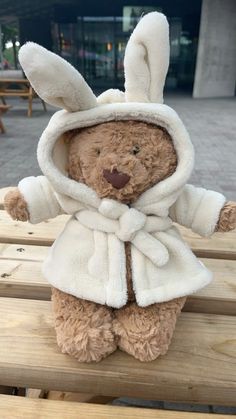 a teddy bear dressed in white sitting on a wooden bench outside with its arms outstretched