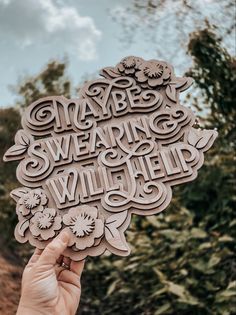 someone holding up a wooden cutout that says maybe swearing and wildflowers