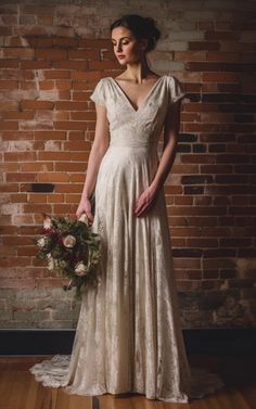 a woman standing in front of a brick wall wearing a dress and holding a bouquet