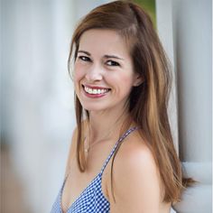 a woman in a blue and white checkered top smiles at the camera while leaning against a column