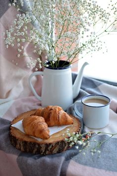 two croissants on a plate next to a cup of coffee and flowers