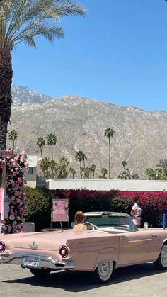 an old pink car is parked in front of a palm tree and some flowers with mountains in the background