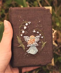 a hand holding up a brown book with a fairy on it's side and flowers in the middle