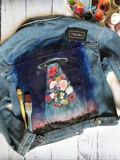 a denim jacket with flowers painted on it and paintbrushes next to the back