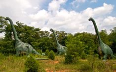 three large dinosaur statues in the middle of a field with trees and bushes behind them