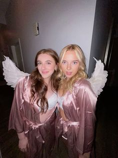 two beautiful young women dressed in pink and white robes posing for the camera with angel wings on their shoulders