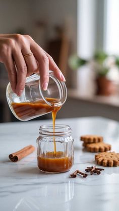 someone pouring caramel sauce into a glass jar with cookies on the table next to it