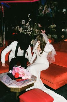 a bride and groom sitting on a red couch in front of a table with flowers