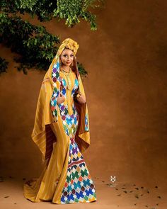 a woman in a yellow dress and head scarf standing under a tree with her hands on her hips