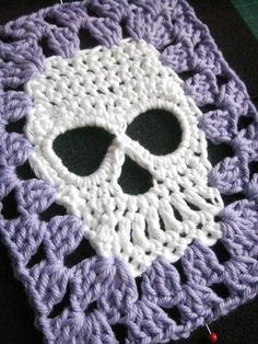 a crocheted square with a white and purple skull in the middle, on a black surface