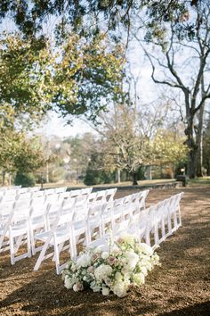 an outdoor ceremony setup with white chairs and flowers on the ground in front of trees