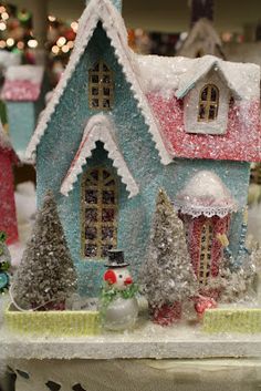 a small blue and red house with snow on it's roof, surrounded by christmas trees