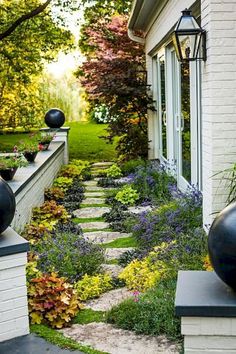 an outdoor garden with flowers and plants on the side of a white brick building, surrounded by greenery