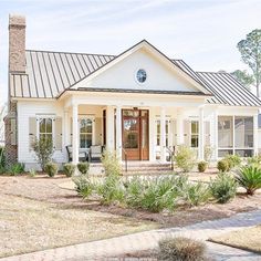 a white house with a metal roof and front porch is surrounded by greenery, shrubs and trees