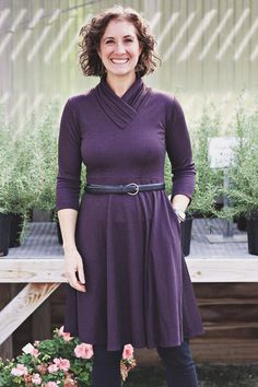 a woman in a purple dress standing next to some potted plants and smiling at the camera