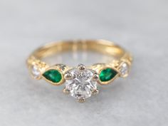 This diamond and emerald engagement is sleek and stylish! The pear-cuts of the emeralds give the ring a contemporary feel, while the ornately textured gold feels luxurious on the hand. Perfect for an engagement or anniversary this is a stunning ring set with a high-quality modern round brilliant cut diamond!  Metal: 14K Yellow and White Gold Gem: Diamond .50 Carats, SI1 in Clarity, G in Color Gem Measurements: 4.8 mm, Round Accents: 2 Emerald totaling .16 Carats, 2 Diamonds totaling.08 Carats  R Diamond And Emerald Engagement Ring, Gold Band Engagement Rings, Emerald Engagement Ring Set, Emerald Wedding Rings, Emerald Ring Gold, Elegant Engagement Rings, Colored Engagement Rings, Yellow Gold Diamond Ring, Diamond Anniversary Rings