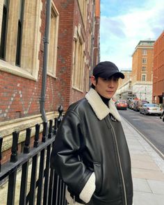 a man standing next to a fence in front of a brick building wearing a black jacket and hat