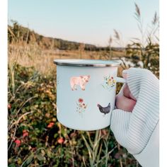 a person holding a cup with animals painted on it in front of some bushes and flowers