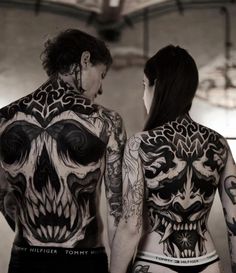 two people with tattoos on their back standing next to each other and looking at each other