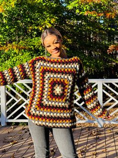 a woman standing in front of a white fence with her arms out and wearing a multicolored crocheted sweater