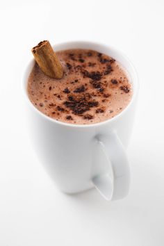 a cup of hot chocolate with cinnamon stick sticking out of it's top, on a white surface