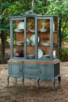 a blue china cabinet with glass doors and plates on it's top, sitting in front of some trees