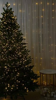 a lit christmas tree in front of a curtained window with lights on it's sides