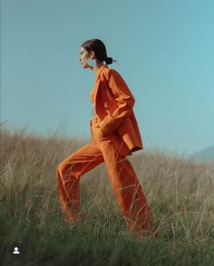 a woman in an orange outfit is walking through tall grass and looking off into the distance