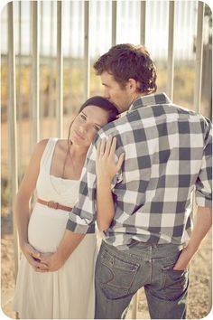 a man and woman standing next to each other in front of a fence with their arms around each other