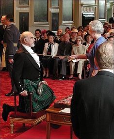 an old man in a kilt is shaking hands with another man who is sitting on a chair