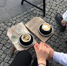 two people sitting at a table with cups of coffee on top of them and one holding the other's hand