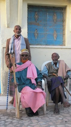 three people sitting on chairs in front of a building with blue doors and windows, one man is wearing a pink scarf