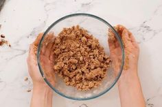 two hands holding a glass bowl filled with granola on top of a marble counter