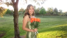 a woman in a green dress is holding some flowers and smiling at the camera while standing under a tree