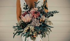 a woman holding a bouquet of flowers and greenery in front of a white wall