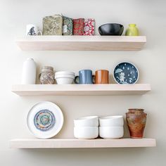 two wooden shelves with bowls, cups and plates on them in a white walled room