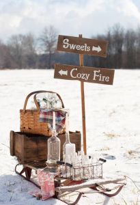 a wooden sign sitting in the snow next to a basket filled with bottles and glasses