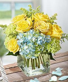 a vase filled with yellow roses and blue hydrangeas on top of a wooden table