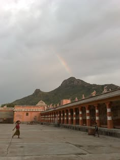 a rainbow is in the sky over a building with arches and pillars, while people are walking around