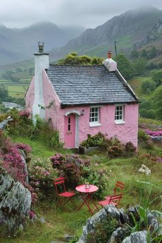 a small pink house sitting on top of a lush green hillside next to flowers and rocks